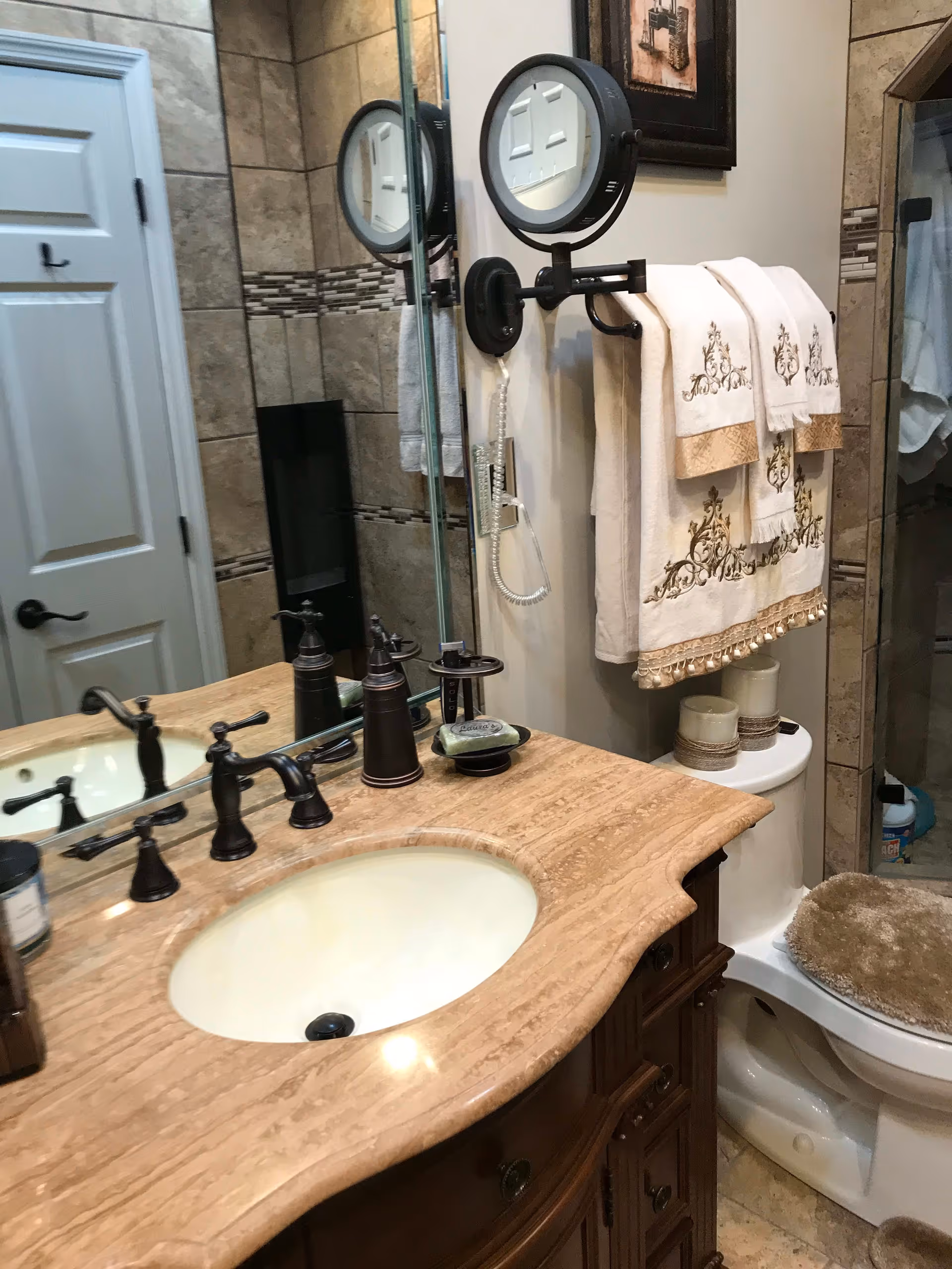 A bathroom with a beige marble countertop and an under-mount sink. The countertop has a bronze faucet, soap dispenser, and soap dish. Above the sink is a large mirror with a mounted round magnifying mirror. On the right side, there are decorative embroidered towels hanging on a towel rack above a toilet with a cushioned seat cover. The walls are tiled with beige tiles and decorative mosaic accents. A white door and a glass shower door are visible in the background.