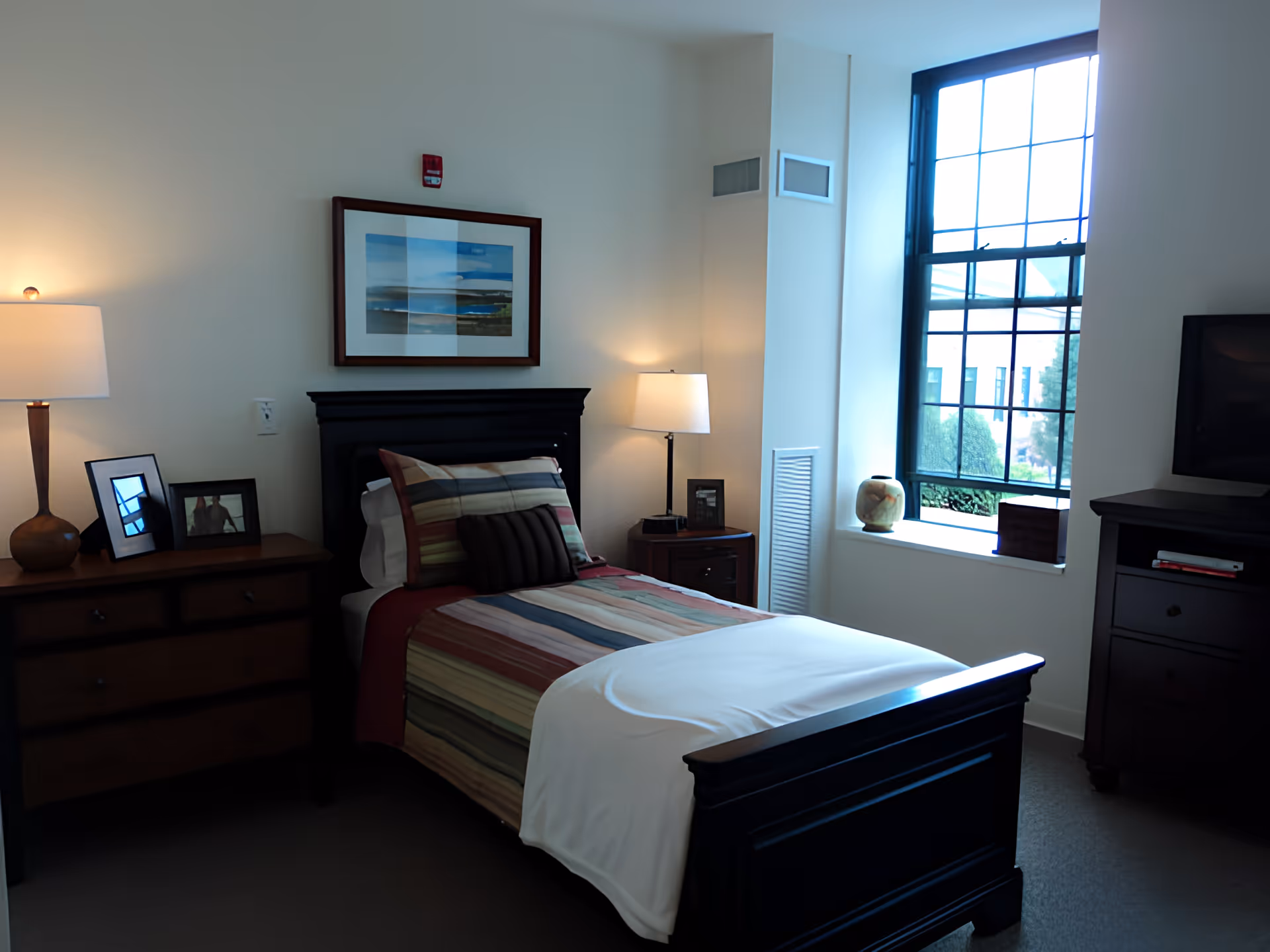 Sunlit bedroom with a single bed dressed in striped bedding, nightstands with lamps, a dresser, and a large window.