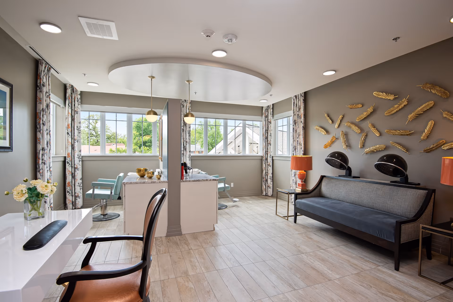 A bright and spacious salon area in a senior living facility with large windows letting in natural light. The room features a modern styling station with chairs, a sofa with two vintage hair dryers, decorative golden feather wall art, and side tables with orange lamps. Light wood flooring and floral curtains add warmth to the space.