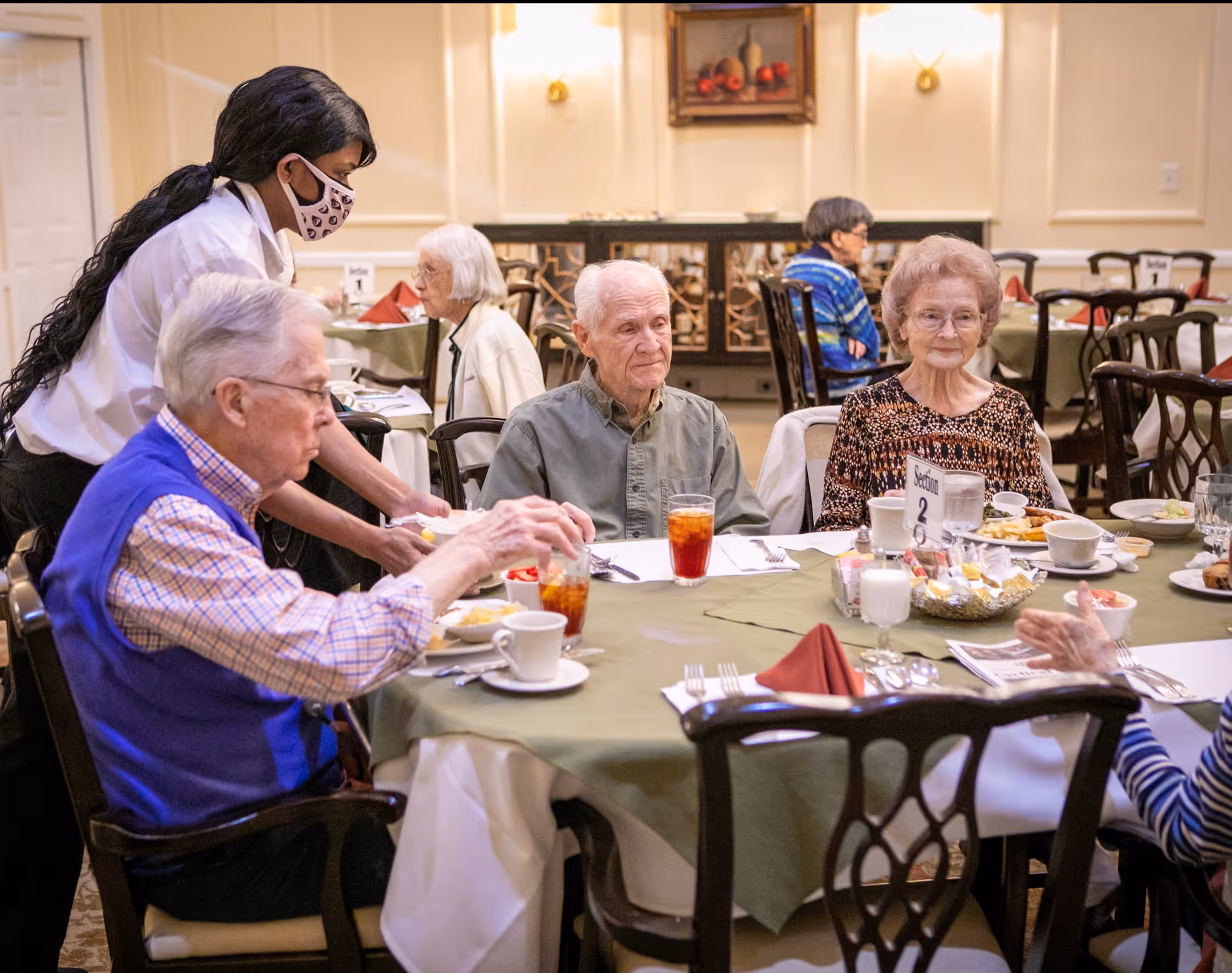 A dining room scene at The Orchard with elderly residents seated around a table set with plates, cups, and glasses of iced tea. A staff member wearing a face mask is serving food to one of the residents. The room has warm lighting, classic wooden chairs, and tables covered with green tablecloths and red napkins.