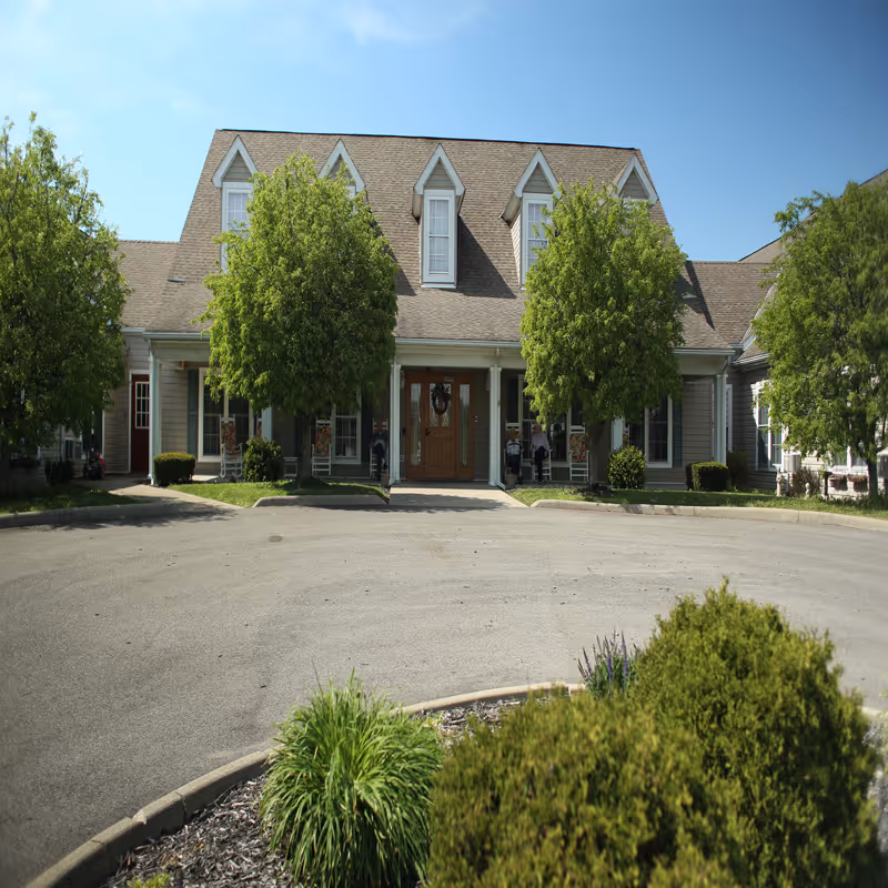 Front exterior view of a senior living facility building with a peaked roof, multiple dormer windows, a central entrance with double wooden doors, and several green trees and shrubs in front under a clear blue sky.