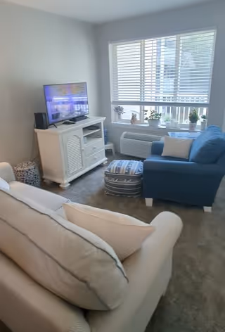A cozy living room with a white couch and a blue armchair facing a white TV stand with a flat-screen television. The room has a window with white blinds, a small ottoman with a patterned cover, and several small potted plants on the windowsill. The floor is carpeted in a neutral color.