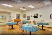 A dining room in a senior living facility with round tables covered in blue tablecloths, set with plates and utensils. Several upholstered chairs are arranged around the tables. The room has light-colored walls decorated with framed pictures and wall-mounted decorations. A piano is visible against one wall.