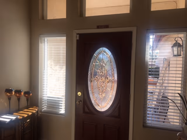 Interior view of an entrance door with an oval decorative glass panel. The door is flanked by two windows with white blinds, and there is a small table with three decorative candle holders on the left side. Above the door is a small exit sign.