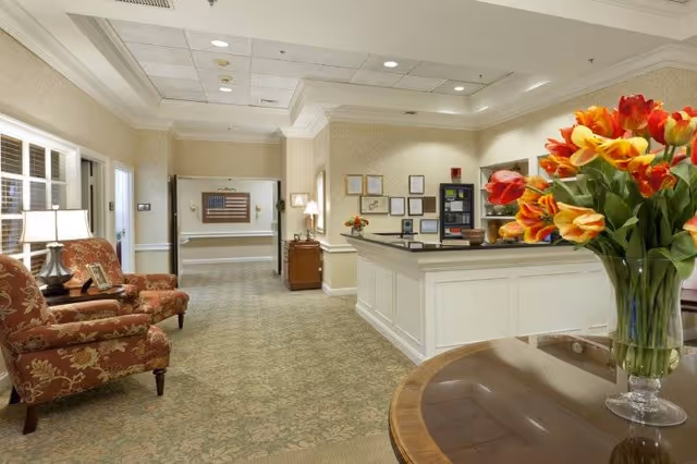 A well-lit reception area in a senior living facility with a white front desk, a vase of red and yellow tulips on a round table, two patterned armchairs with a side table and lamp, and framed certificates on the wall behind the desk. The hallway extends into the background with an American flag displayed on the far wall.