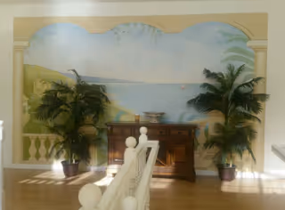 Interior view of a room with a large mural depicting a seaside landscape with a sailboat on the water. In front of the mural is a wooden sideboard with decorative items on top, flanked by two large potted palm plants. The room has light-colored walls and wooden flooring, with a white railing in the foreground.