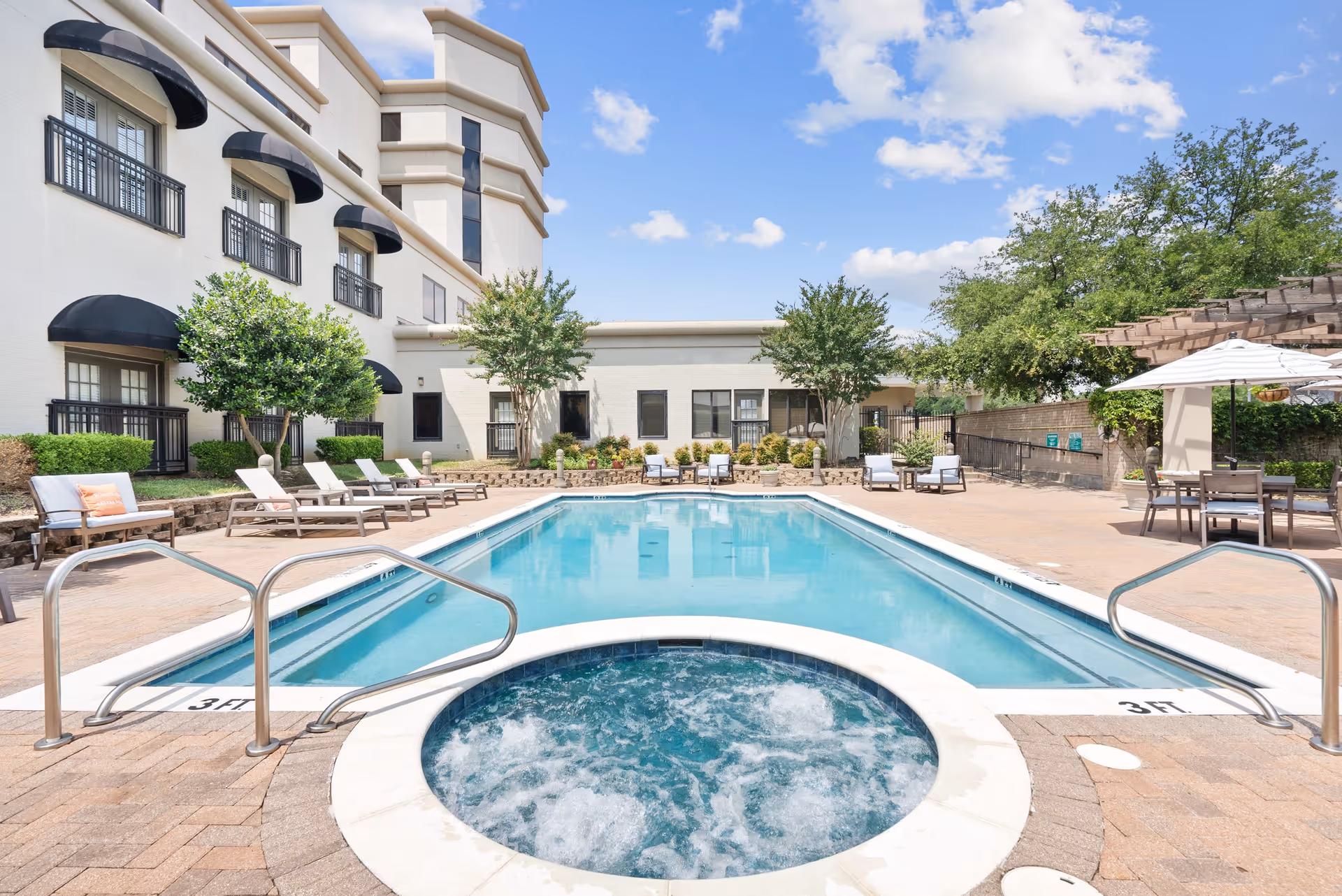 Outdoor swimming pool area at Parc Place with a rectangular pool and a circular hot tub in the foreground. The pool is surrounded by lounge chairs, tables with umbrellas, and greenery including trees and shrubs. The building with black awnings and windows is visible in the background under a partly cloudy blue sky.