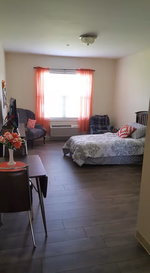 A bright bedroom with a large window covered by sheer orange curtains. The room features a bed with patterned bedding and a red and white decorative pillow, two upholstered armchairs with cushions, a small table with a vase of flowers, and a wall-mounted TV. The floor is wood, and the walls are painted light beige.