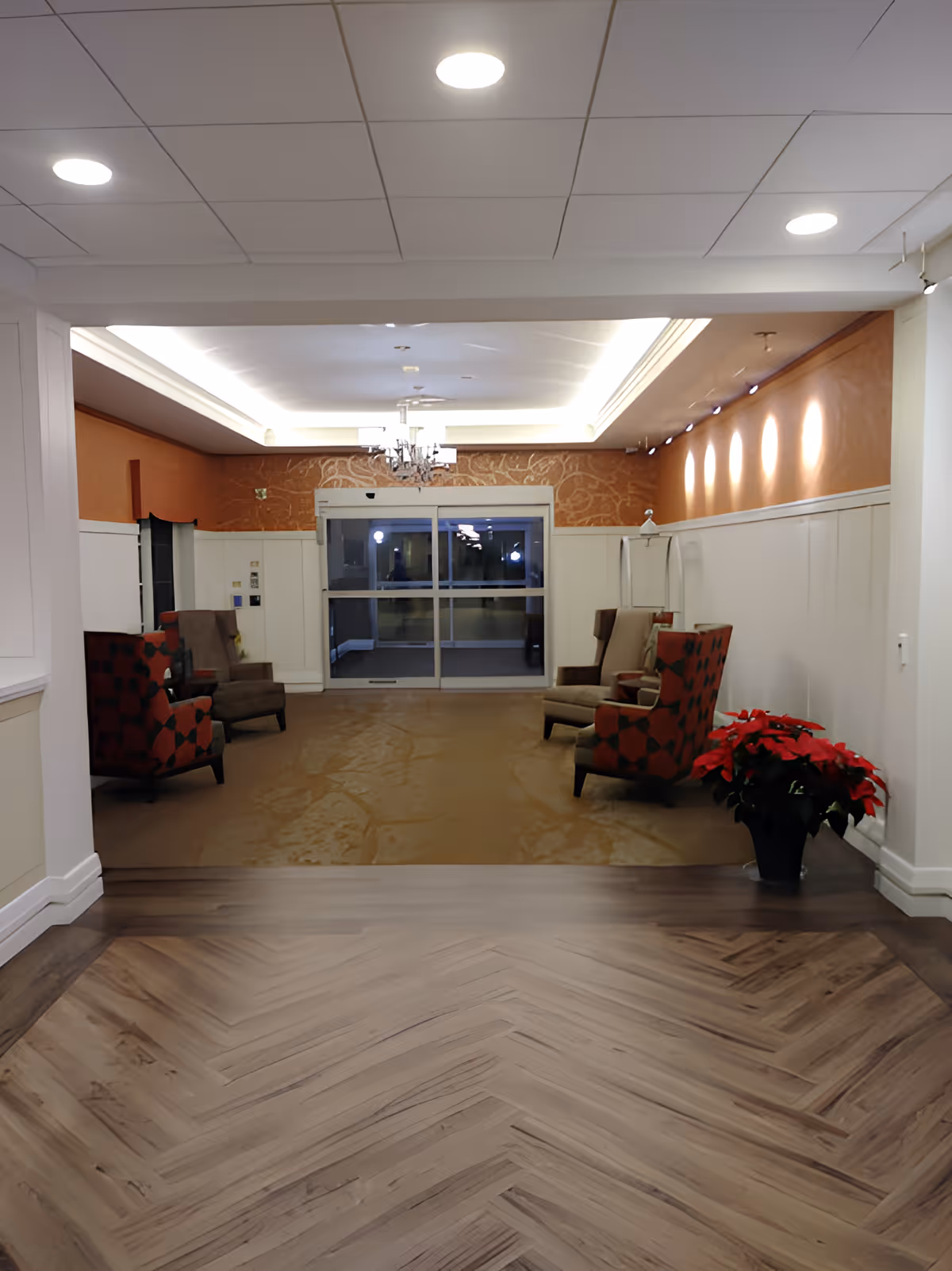 A well-lit interior lounge area with four armchairs arranged in pairs along the walls. The walls are painted white with an orange accent wall at the far end, decorated with a subtle pattern. There is a chandelier hanging from the ceiling and recessed lighting along the right wall. A potted poinsettia plant is placed on the right side near the entrance. The floor transitions from a wood-patterned surface in the foreground to a carpeted area in the seating space. Glass double doors are visible at the far end of the room.
