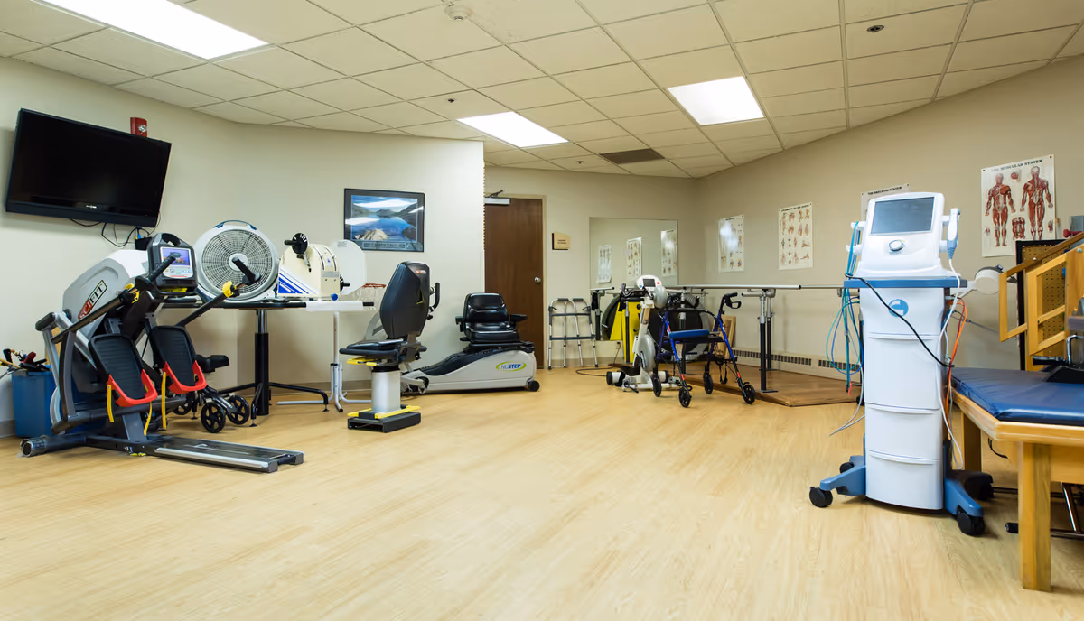 A physical therapy room in an assisted living facility featuring exercise equipment including a recumbent bike, a NuStep machine, walkers, parallel bars for walking support, and a medical device on wheels. The room has light wood flooring, beige walls with anatomical charts, a wall-mounted TV, and a large mirror.