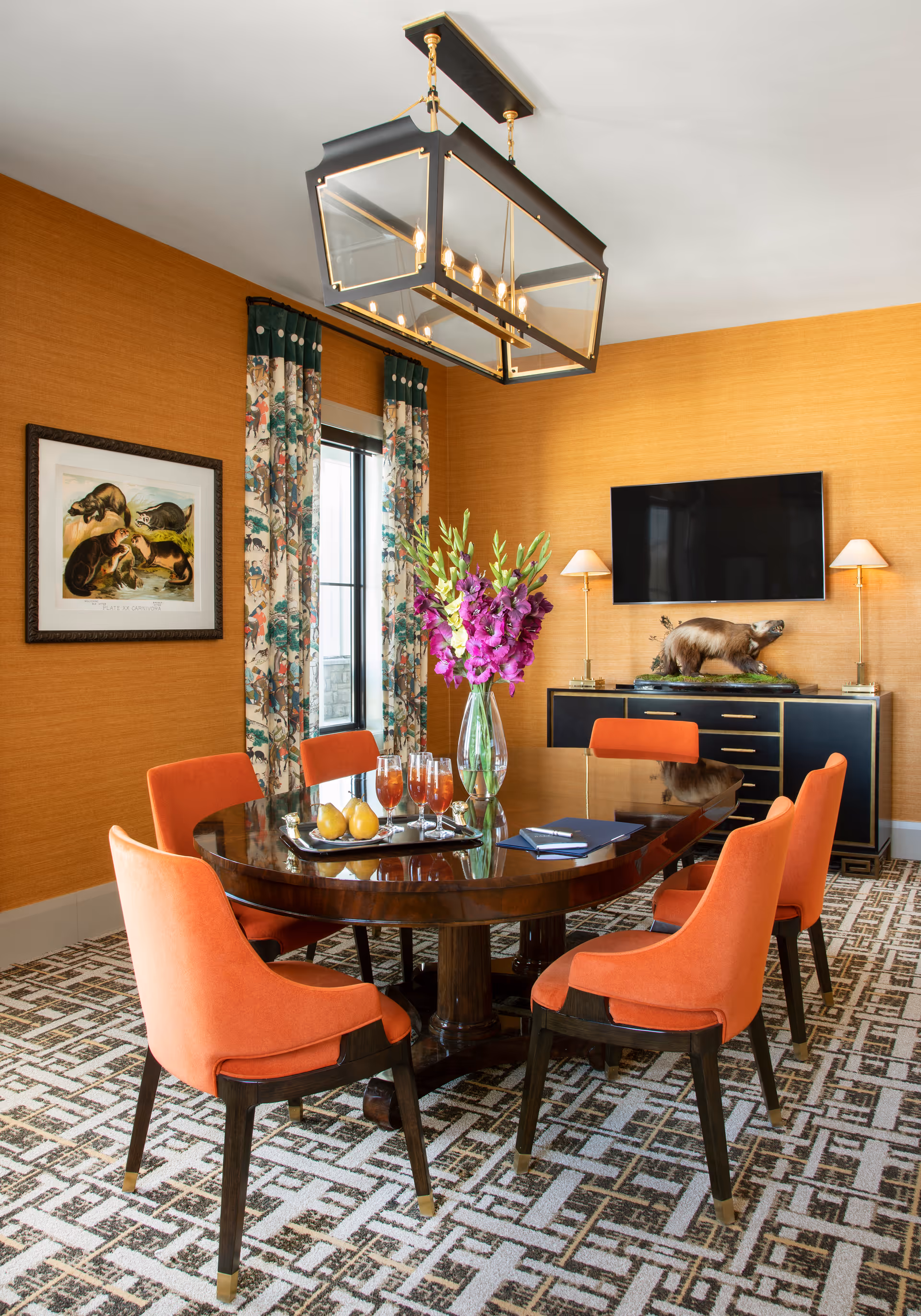 A dining room with a dark wooden oval table surrounded by six orange upholstered chairs. On the table is a vase with purple flowers, a tray with drinks and pears, and a closed notebook. The room has orange textured walls, patterned curtains with a nature motif, a framed animal artwork, a patterned carpet, and a black sideboard with gold accents. Above the sideboard is a mounted flat-screen TV and two wall lamps. A large rectangular chandelier hangs from the ceiling.