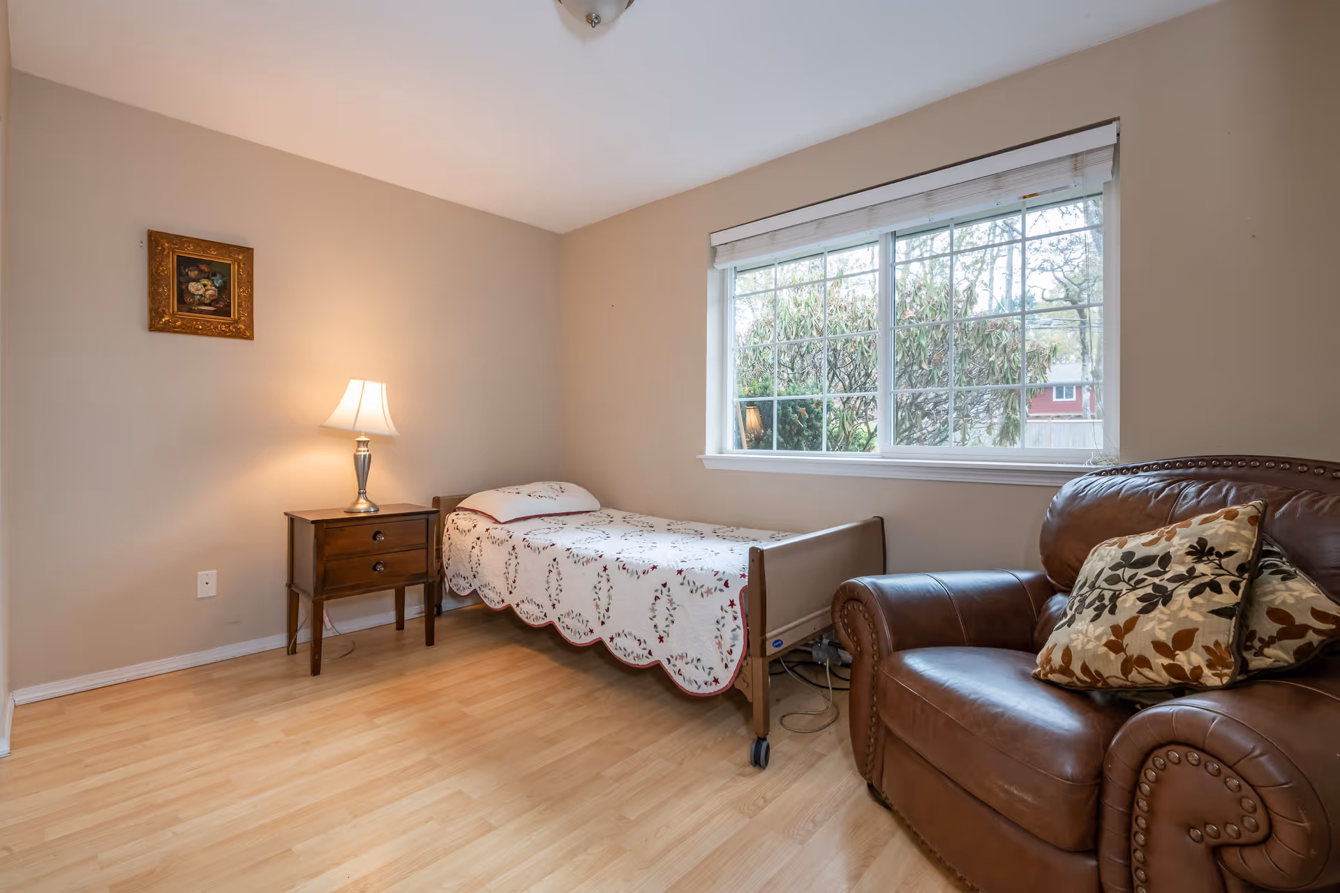 A small bedroom with a single bed covered with a floral quilt, a wooden nightstand with a lamp, a large window with a view of trees outside, and a brown leather armchair with two decorative pillows.