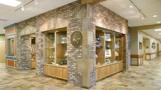 Interior hallway corner of a facility with brick and tile walls featuring glass display cases containing various memorabilia and artifacts. The floor is tiled, and the ceiling has recessed lighting. A seal or emblem is mounted on the brick wall at the corner.