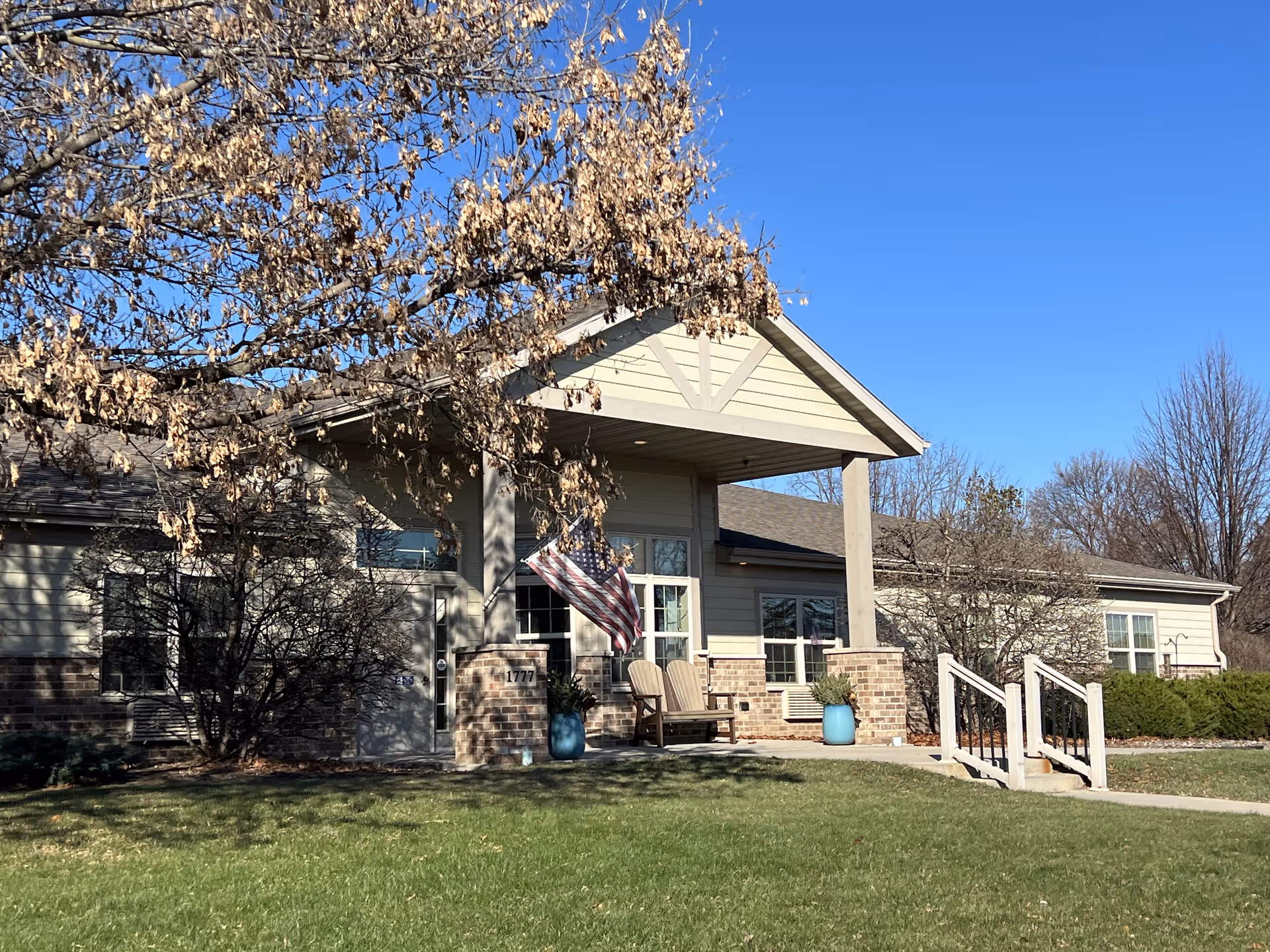 Exterior view of a single-story building with a covered entrance supported by two pillars. There are two wooden chairs and two blue planters near the entrance. An American flag is hanging near the door. The building is surrounded by grass, leafless trees, and shrubs under a clear blue sky.