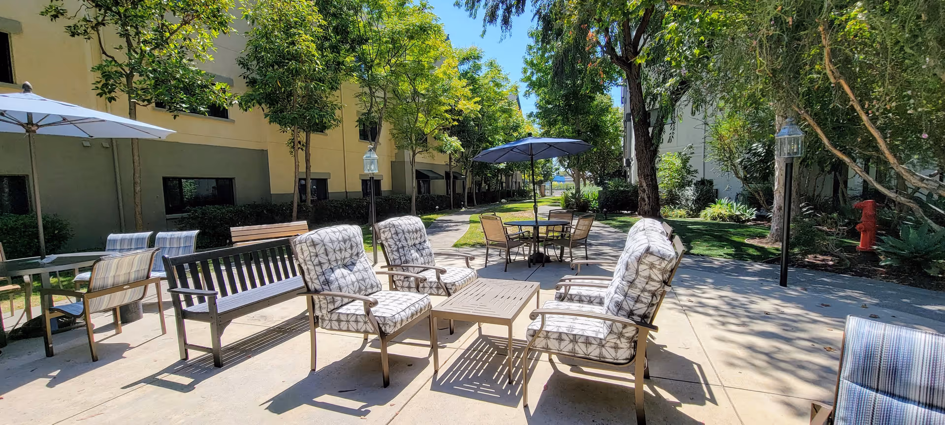 Outdoor patio area at Westminster Terrace Senior Living with cushioned chairs, a bench, tables with umbrellas, surrounded by trees and greenery on a sunny day.