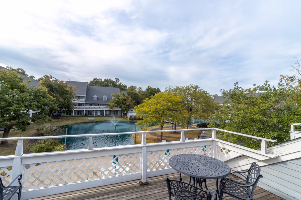 View from a balcony with table and chairs overlooking a pond with a fountain, trees, and surrounding senior living buildings.