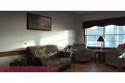 A small sitting room with an upholstered sofa, armchairs, side tables and a lamp in front of a large window.
