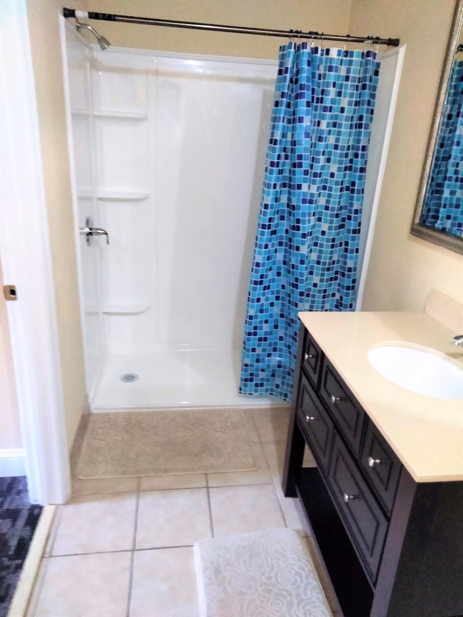 Bathroom with a white tiled floor, a walk-in shower with a blue and turquoise mosaic patterned shower curtain, a beige bath mat in front of the shower, and a dark wood vanity with a beige countertop and an oval sink. A mirror is partially visible on the right wall.