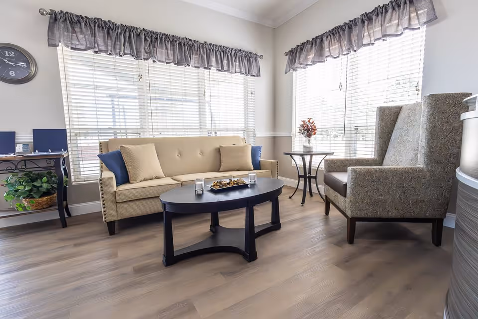 A bright and cozy living room area with a beige sofa adorned with blue and beige cushions, a patterned armchair, a black oval coffee table with decorative items, and two side tables. The room has large windows with white blinds and gray valances, light wood flooring, and a wall clock above a small table with a potted plant.