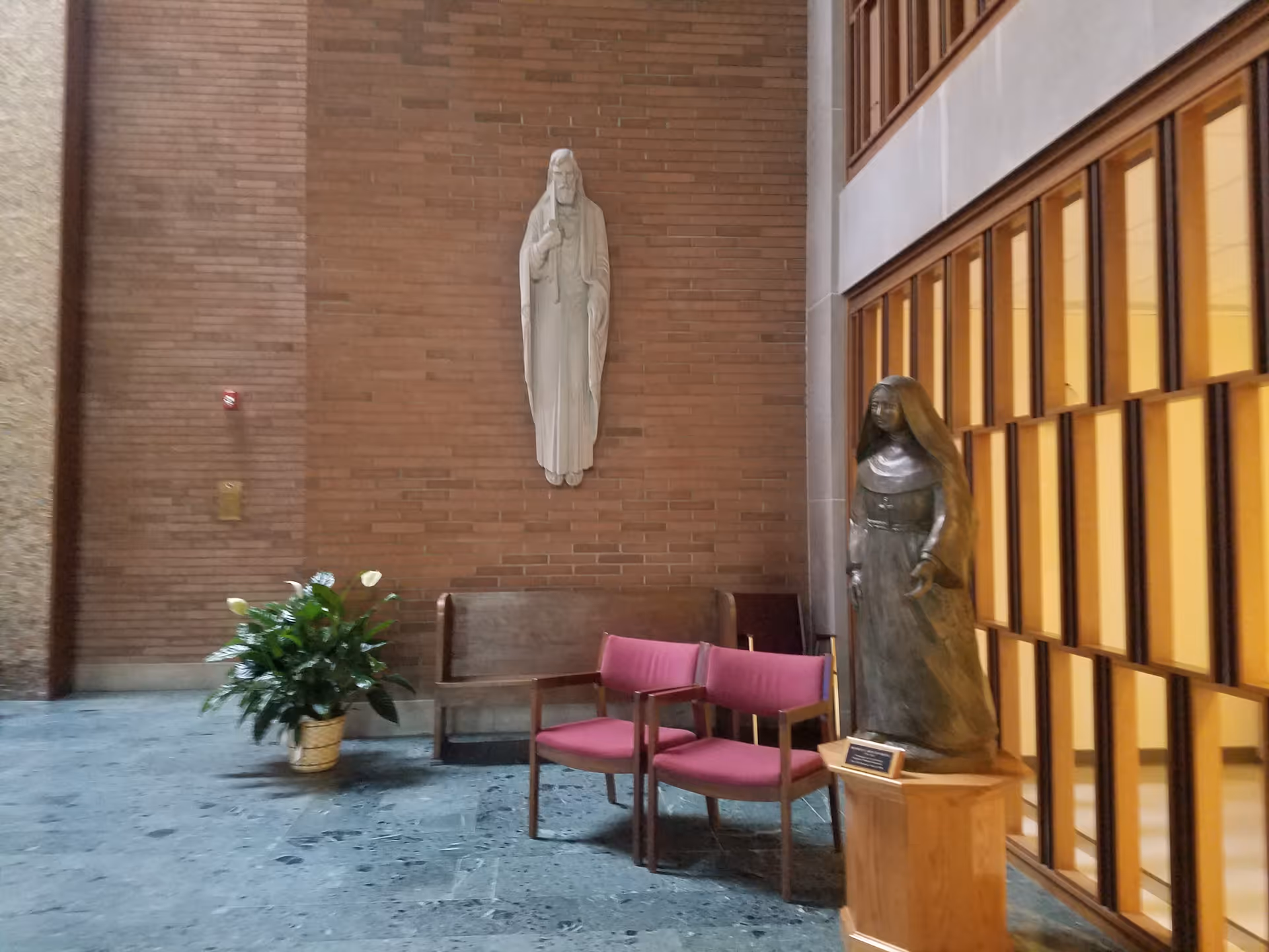 Indoor seating area in a brick-walled chapel or lobby featuring two religious statues, a wooden bench, two pink chairs, and a potted plant.