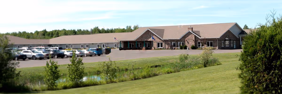Exterior view of a single-story senior living facility building with a stone facade and multiple windows, a parking lot with several cars, a grassy area with small trees and shrubs, and a clear blue sky.