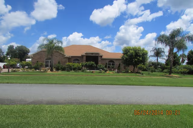 Front exterior view of a single-story brick building with a brown roof, surrounded by green grass, palm trees, and shrubs under a partly cloudy blue sky.