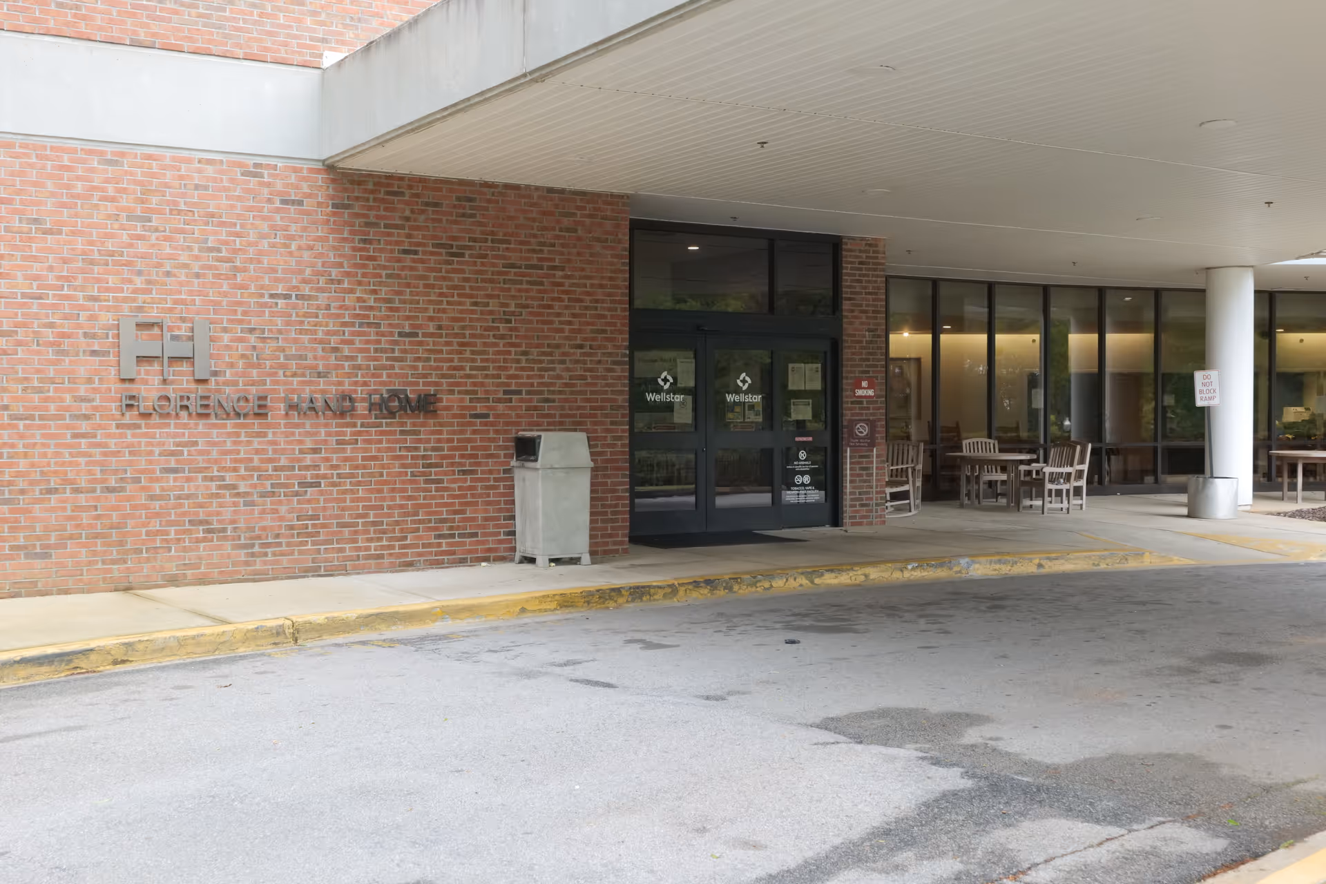 Entrance of Florence Hand Home facility showing a brick wall with the facility name and logo, glass double doors with Wellstar signage, and an outdoor seating area with tables and chairs under a covered drop-off area.