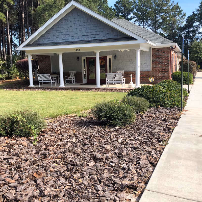 Front exterior view of a single-story building with a covered porch supported by white columns. The porch has white rocking chairs and benches. The building has a gray shingled gable roof and brick walls. There is a well-maintained lawn and landscaped bushes in front, with a concrete sidewalk on the right side.