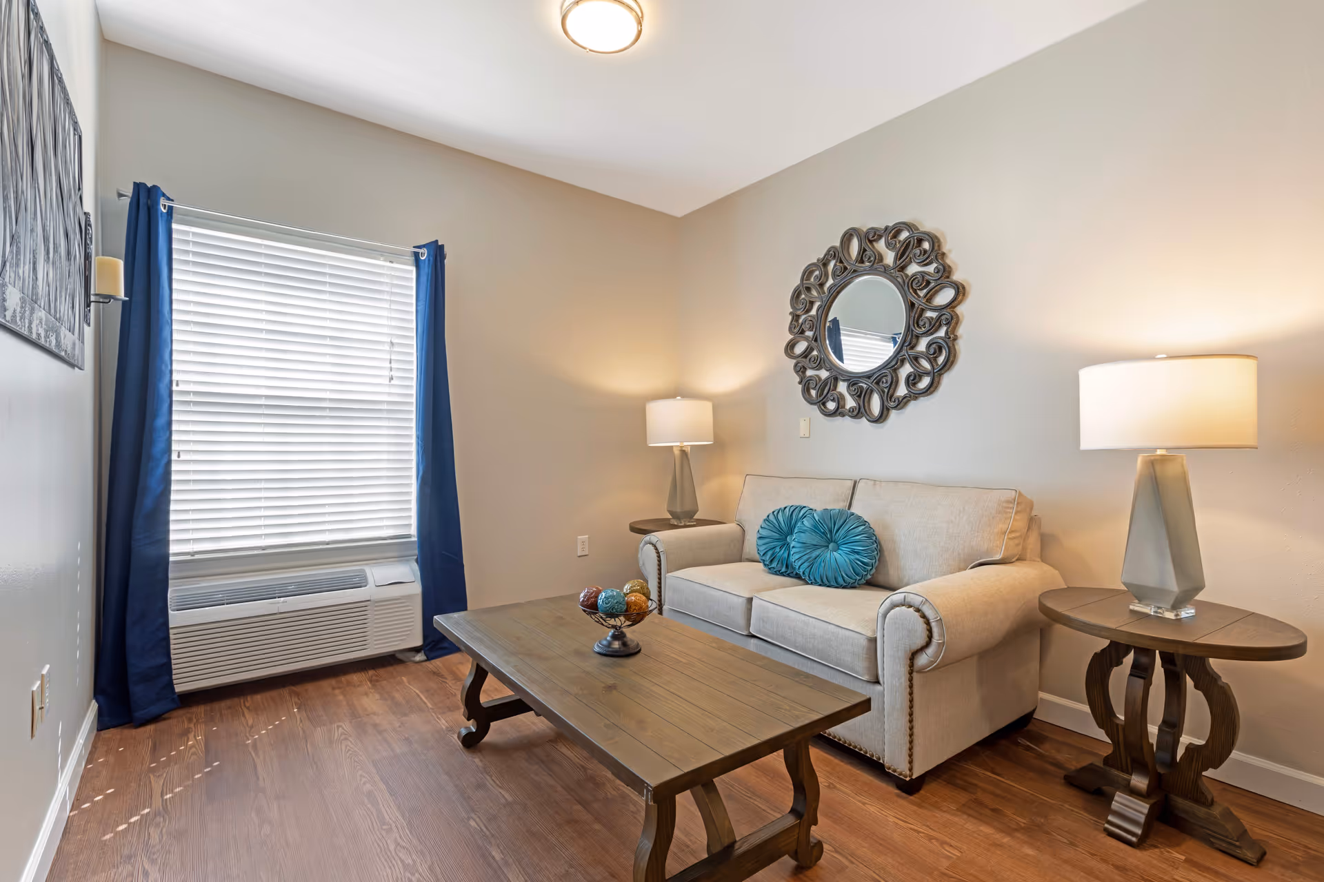 A cozy living room with a beige sofa adorned with two round teal pillows, a wooden coffee table with decorative balls, two side tables each with a lamp, a decorative round mirror on the wall, and a window with blue curtains and white blinds. The floor is wooden and the walls are painted light beige.
