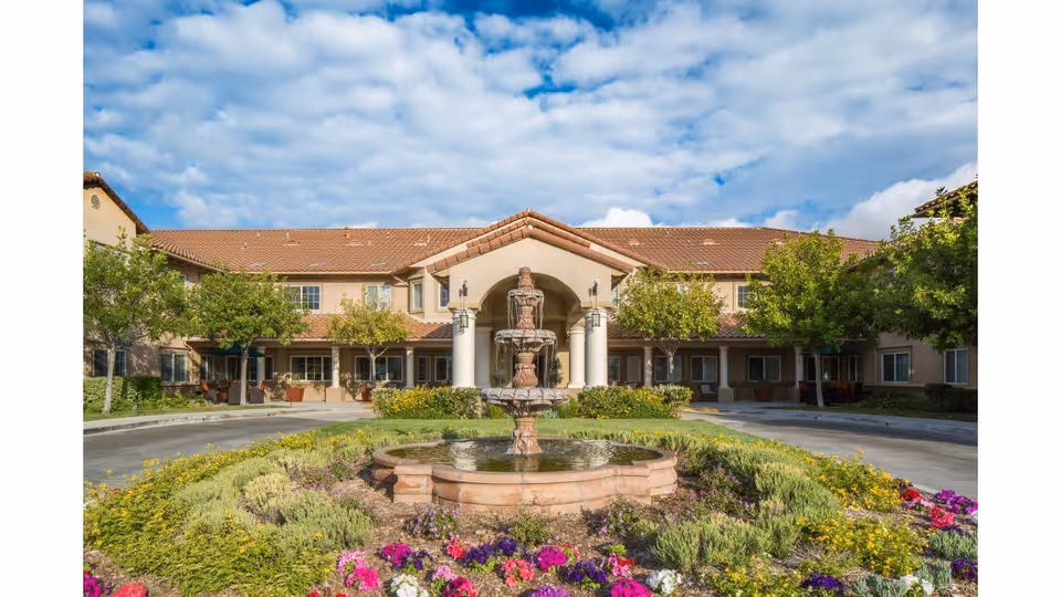 Front exterior of a two-story senior living facility with a central fountain, landscaped flowerbeds, and a tiled roof under a partly cloudy sky.