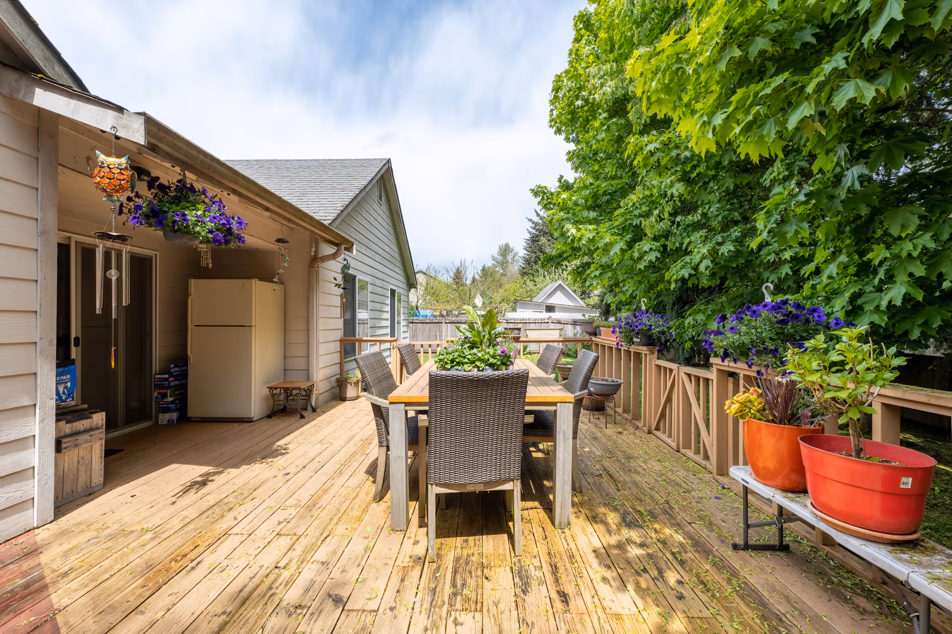 Outdoor wooden deck area with a dining table and six wicker chairs. The deck is decorated with potted plants and hanging flower baskets. There is a white refrigerator against the house wall, and a wind chime hanging from the roof. Trees and greenery surround the deck under a partly cloudy sky.