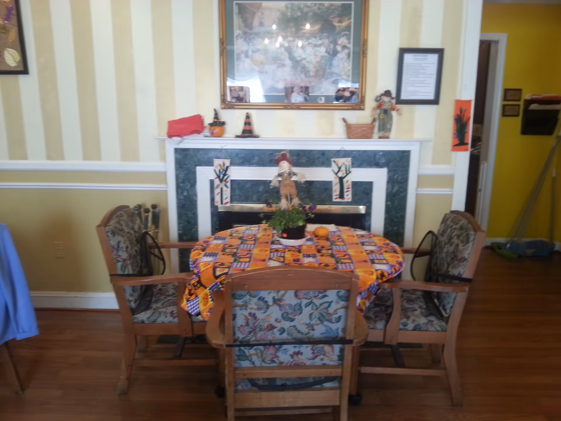 A cozy dining area with a round table covered by a colorful Halloween-themed tablecloth. Four wooden chairs with floral upholstery surround the table. Behind the table is a fireplace with a green marble surround and a white mantel decorated with small Halloween decorations, a basket, and framed pictures. The walls have yellow and cream vertical stripes, and there is a framed picture hanging above the fireplace.