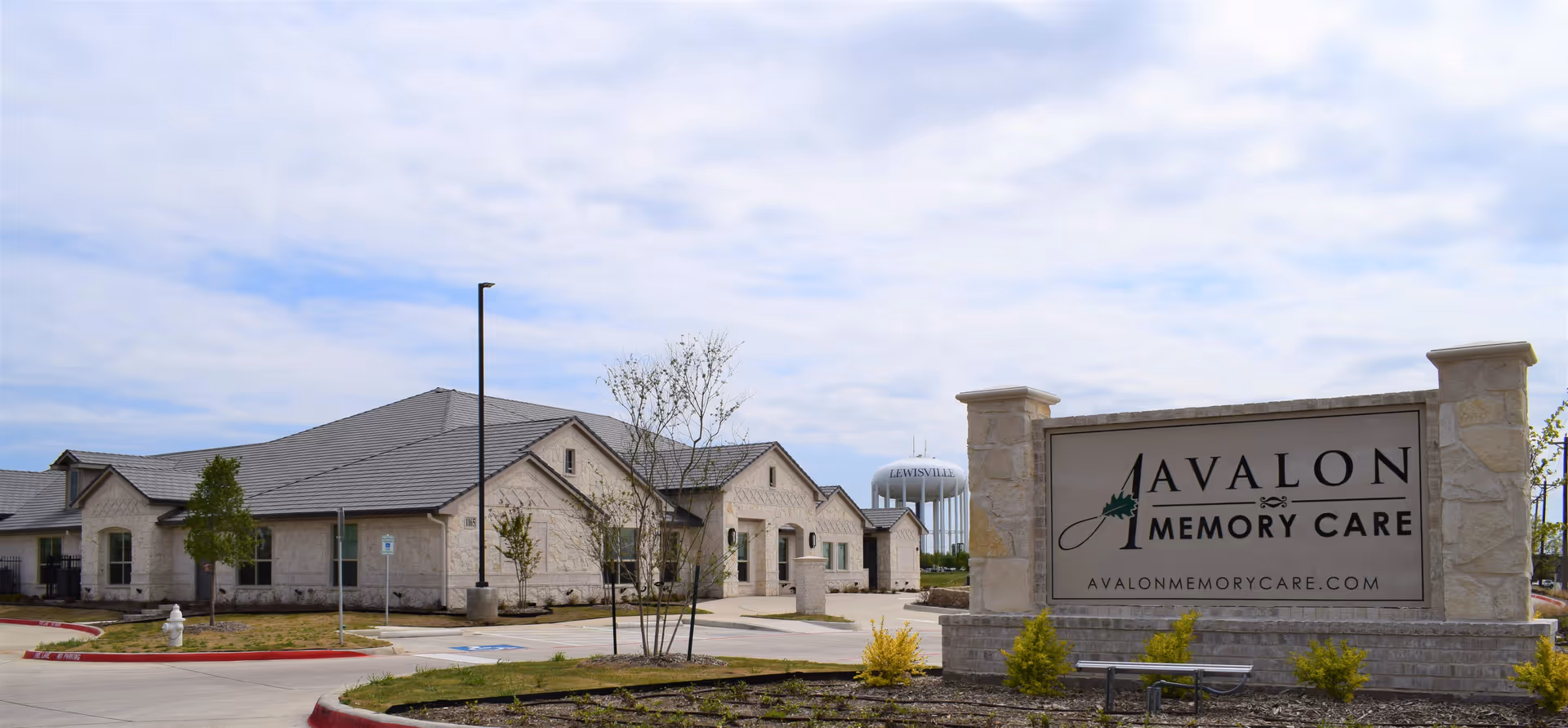 Exterior view of Avalon Memory Care facility with a large stone sign displaying the facility name and website. The building is a single-story structure with a gray roof and light-colored stone walls. There is a water tower in the background labeled 'Lewisville'. The sky is partly cloudy.