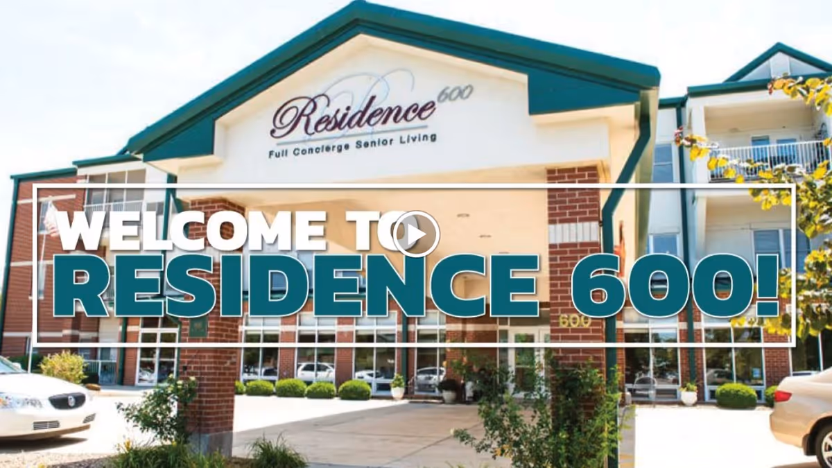 Front exterior view of Residence 600, a senior living facility with a covered entrance, brick and beige walls, green roof accents, and landscaping including bushes and trees. Two cars are parked in front of the building.