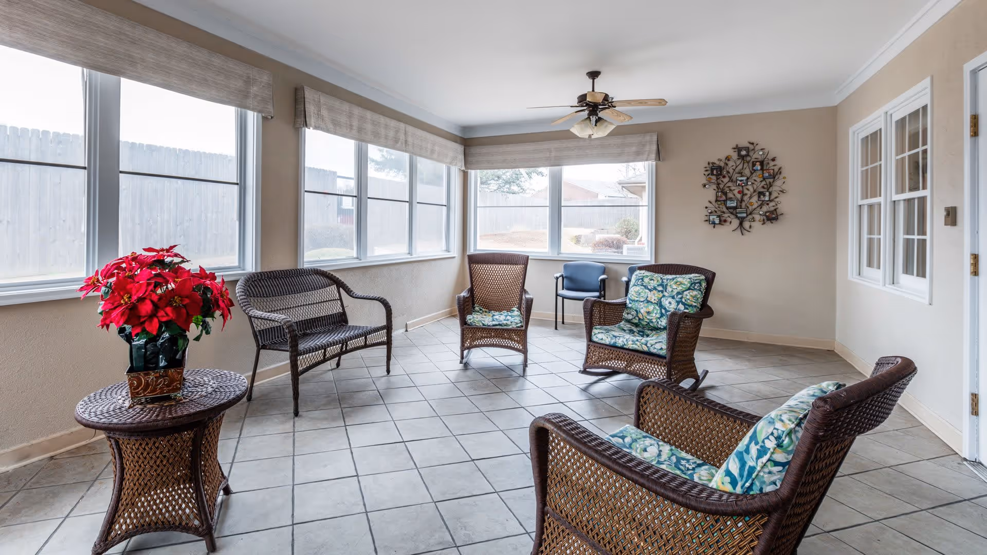 A bright sunroom with large windows on two walls, featuring wicker furniture including chairs and a loveseat with floral cushions, a wicker side table with a red poinsettia plant, a ceiling fan, and a decorative wall piece shaped like a tree with photo frames.