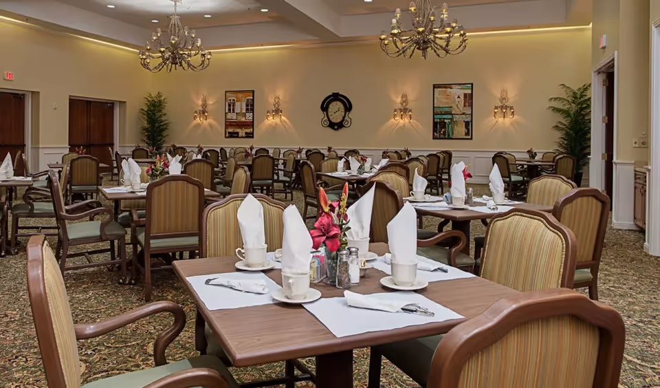 A spacious dining room with multiple tables set for a meal, each table featuring white napkins folded upright, cups, and floral centerpieces. The room has elegant chandeliers, wall sconces, framed artwork, and potted plants, with carpeted flooring and wooden chairs with cushioned seats.