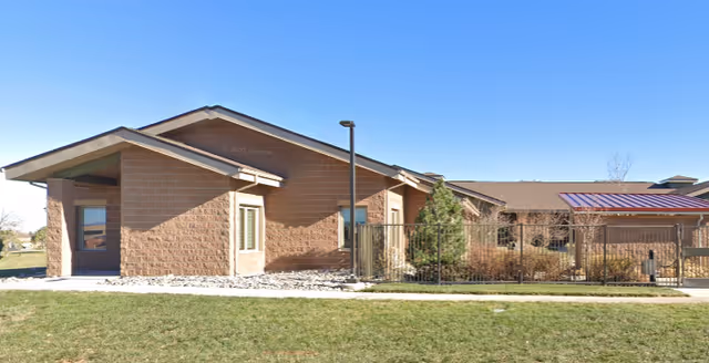 Exterior view of a single-story brick building with a sloped roof under a clear blue sky. The building has several windows and a fenced area with some shrubs and small trees. There is a grassy lawn in the foreground.