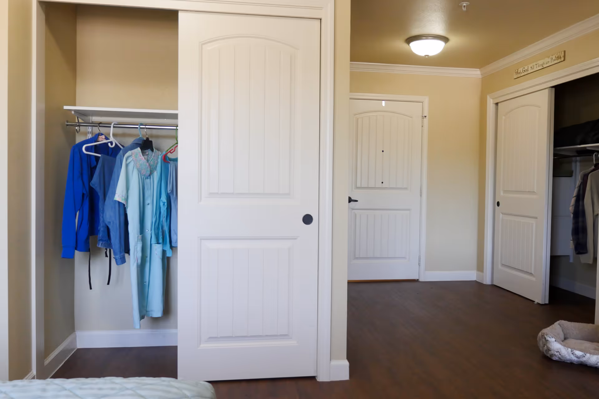Interior view of a room with two open closets showing hanging clothes. The room has beige walls, white doors, and wood flooring. A pet bed is visible on the floor near one of the closets.