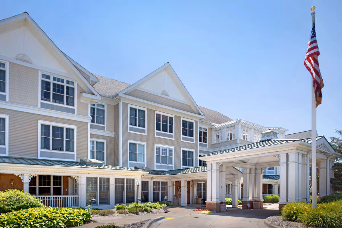 Exterior view of a multi-story senior living facility with beige siding and white trim. The building has multiple windows and a covered entrance with white columns. An American flag is flying on a flagpole near the entrance, and there are green shrubs and plants around the driveway.