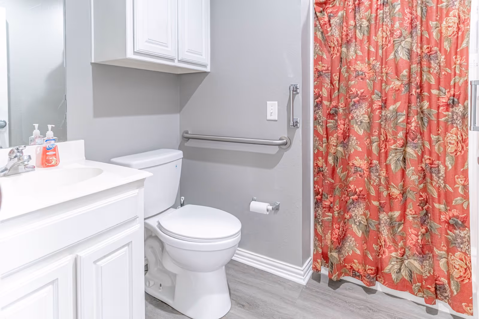 A clean bathroom featuring a white toilet, a white vanity with a sink and faucet, a mirror above the sink, a wall-mounted cabinet, a grab bar next to the toilet, a toilet paper holder, and a shower with a red floral-patterned curtain.