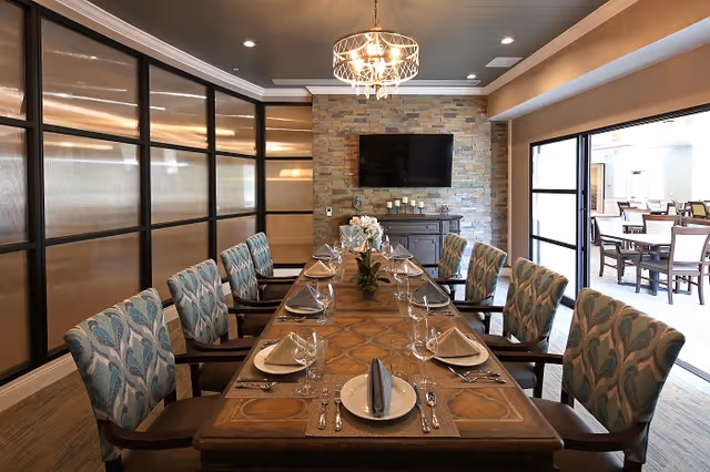 A well-lit dining room with a long wooden table set for eight people. Each place setting includes a plate, folded napkin, glass, and silverware. The chairs have patterned upholstery. A chandelier hangs above the table, and a flat-screen TV is mounted on a stone accent wall at the far end. Large windows and a sliding door open to an adjacent dining area with additional tables and chairs.