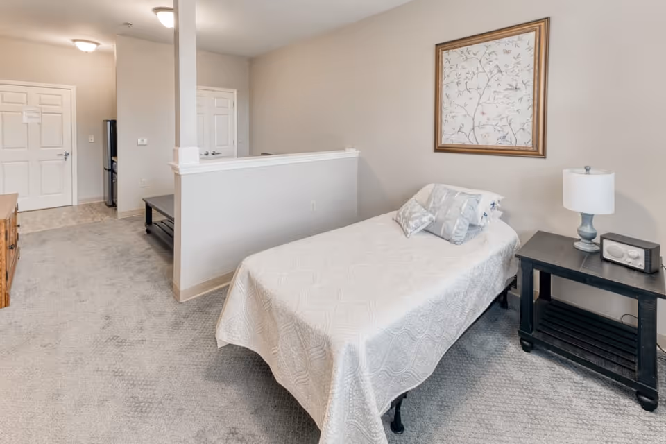 A small bedroom with a single bed covered in a white quilt and two pillows. Next to the bed is a black nightstand with a lamp and a small radio. A framed artwork hangs on the beige wall above the bed. The room has light gray carpet and a partial wall separating the sleeping area from a hallway leading to a door and a kitchen area with a refrigerator.