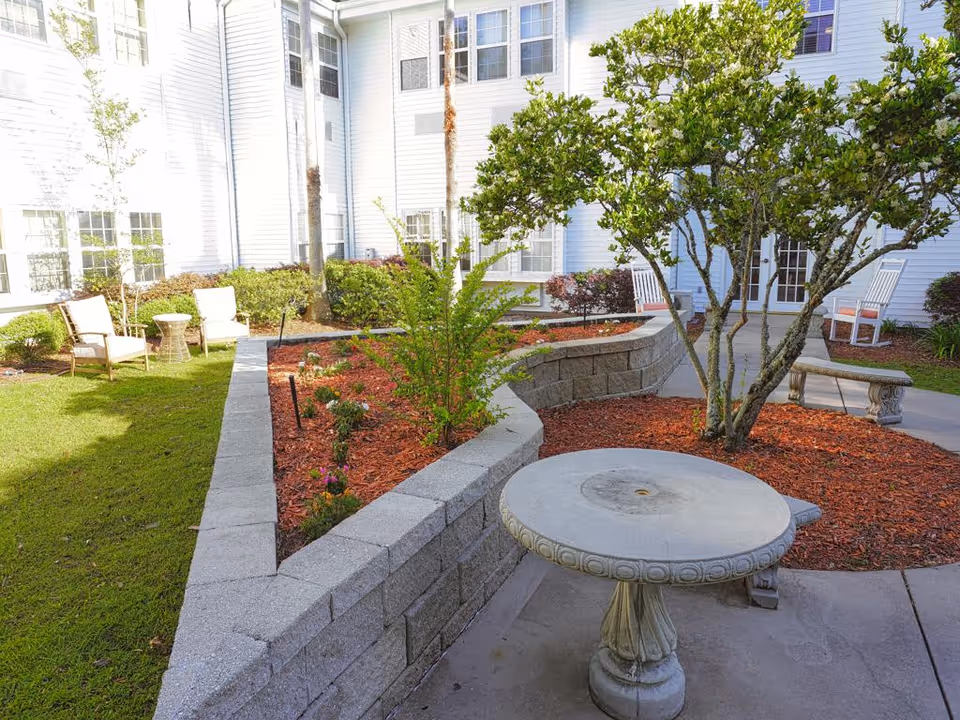 Outdoor courtyard area with a circular stone table and benches, surrounded by landscaped garden beds with mulch and small plants. There are white chairs and a small table on the grass near the building, which has white siding and multiple windows.