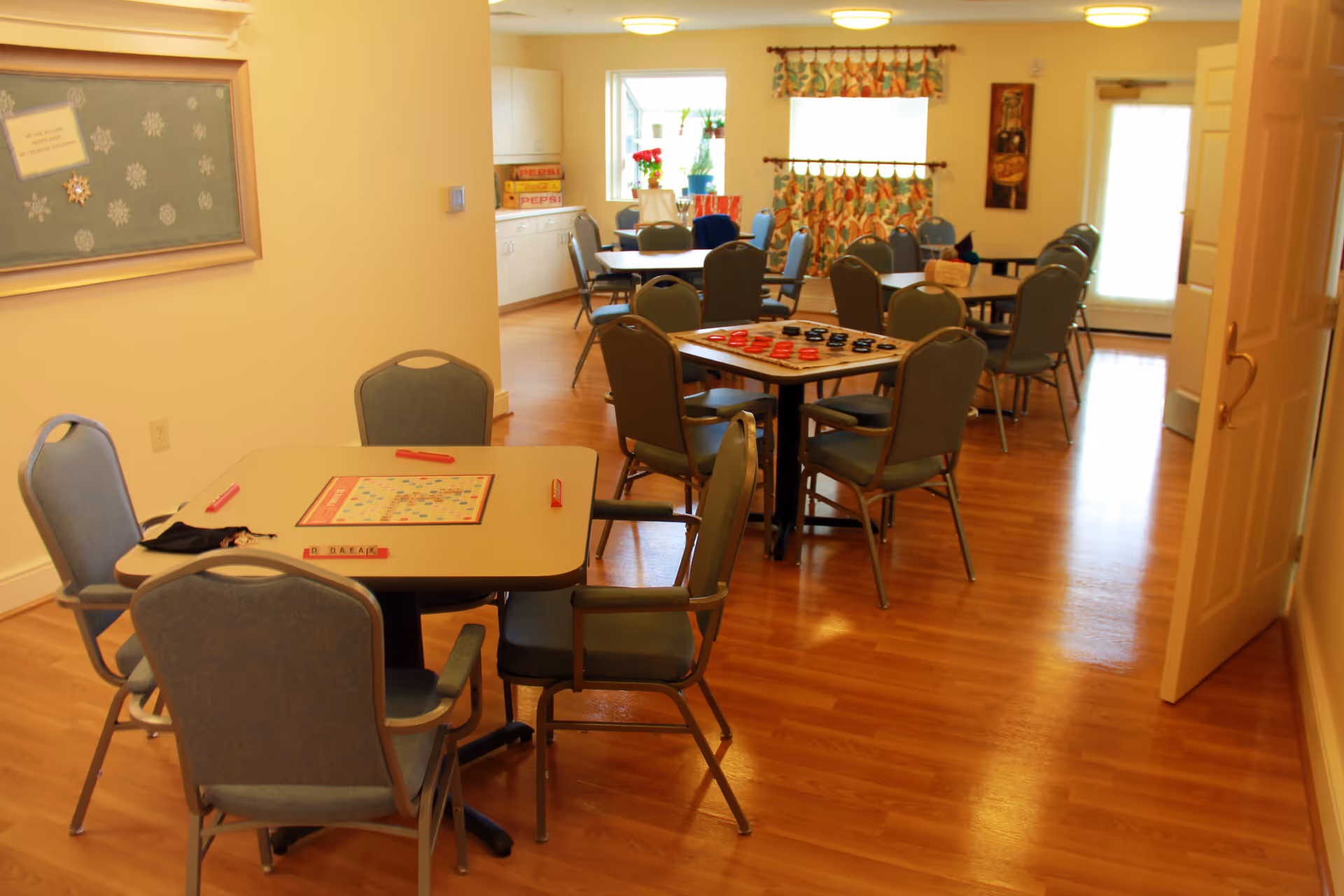 A bright room with several tables and chairs arranged for group activities. One table has a Scrabble board game set up, and another table has a checkers game. The room has wooden flooring, light-colored walls, a bulletin board with snowflake decorations, and windows with colorful curtains letting in natural light.