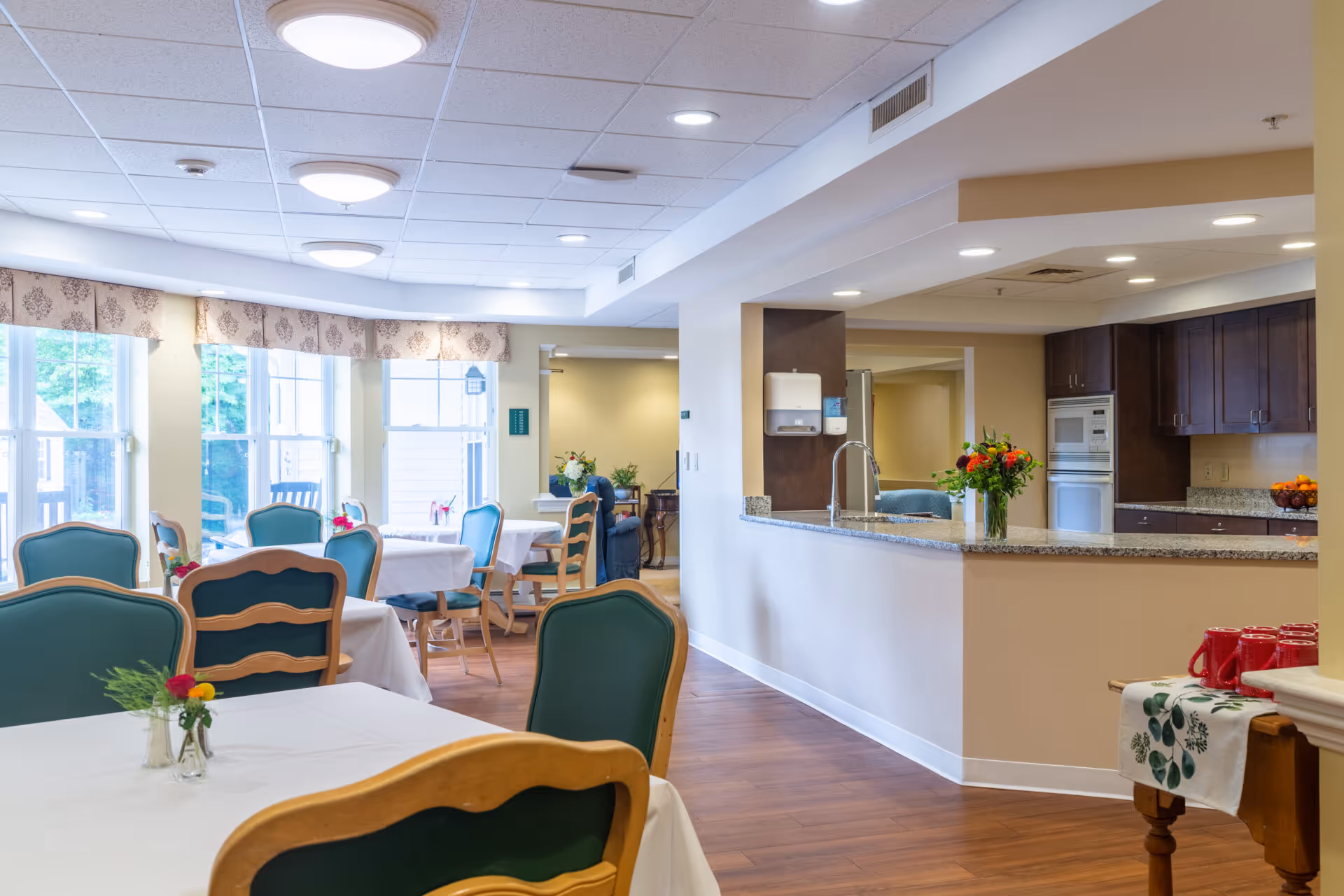 Bright dining area with tables and green-upholstered chairs next to a serving counter and kitchenette.