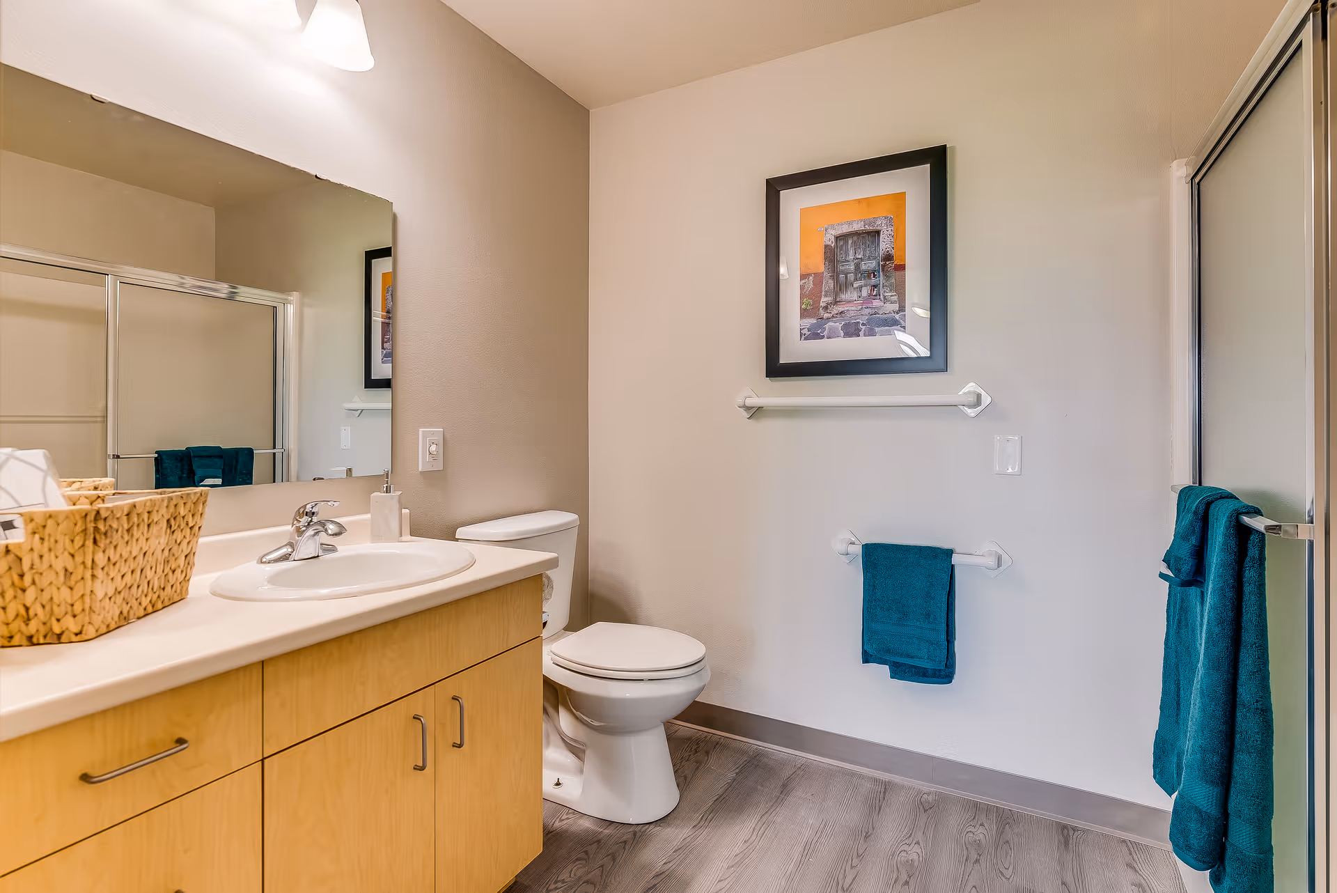 Bright bathroom with a sink vanity, toilet, framed artwork, teal towels, and a glass shower door.