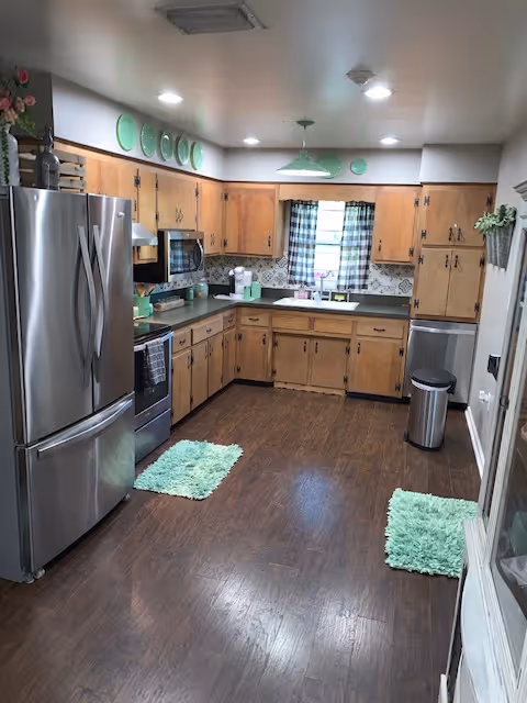 Kitchen with wooden cabinets, stainless steel refrigerator and stove, dark wood floor, and green accent rugs and decor.
