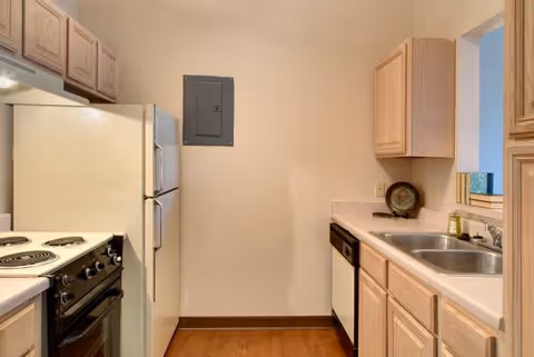 Small kitchen with light wood cabinets, a white refrigerator, an electric stove with coil burners, a dishwasher, a double sink, and a small window opening to another room. The walls are beige and the floor is wood.