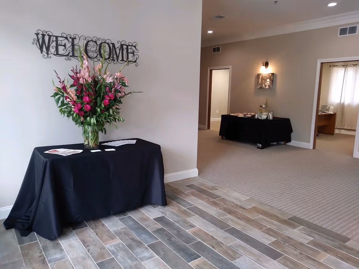 An interior view of a senior living facility entrance area with a black table covered with a black cloth holding a large vase of pink and purple flowers. Above the table is a decorative metal wall sign that reads 'WELCOME'. In the background, there is another black table with refreshments and two open doorways leading to other rooms.