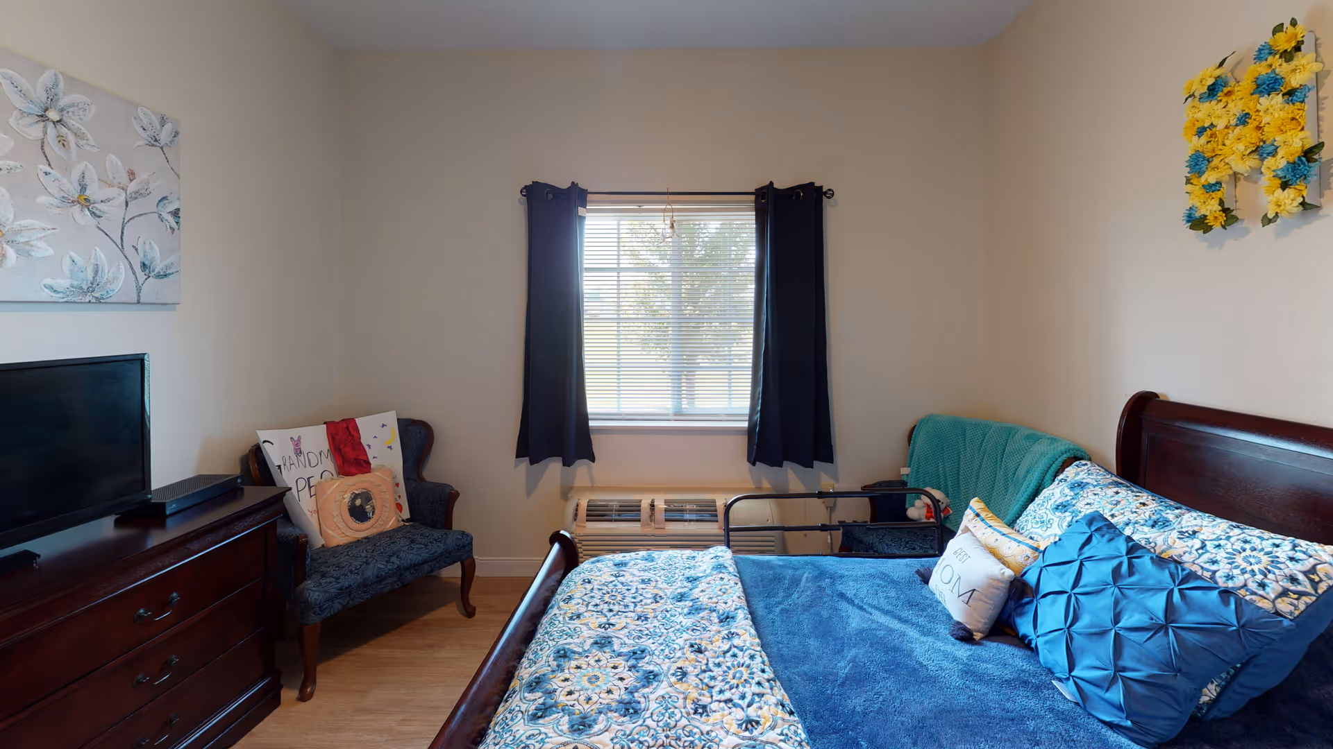 A cozy senior living bedroom with a wooden bed featuring blue and floral bedding, a teal blanket, and decorative pillows. There is a window with dark curtains letting in natural light, a wooden dresser with a flat-screen TV on top, and a cushioned chair with pillows in the corner. The walls are light-colored with floral artwork and a decorative yellow and blue flower arrangement.